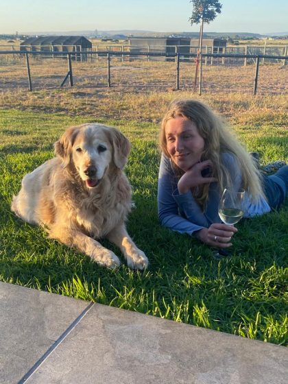 Lilley is lying on her stomach on the grass with her guide dog Teska beside her. She is wearing blue jeans and a long sleeved blue V-necked T-shirt. Her chin rests in her left hand, and she is holding a glass of wine in the other. A stable is visible in the background.