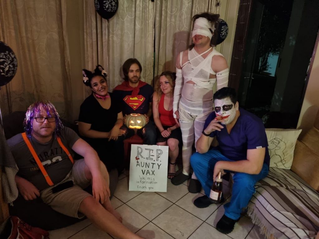 Lilley and a group of friends sit on and stand around a sofa in Halloween costumes. Lilley is dressed as Harley Quin, with a short black ballerina skirt, a red halter top, hair in two long ponytails, and a symbol drawn on her face. There is also a man in a Joker costume, a man wearing a shiny colourful wig, a woman dressed as a black cat, a man dressed as Superman, and a man wrapped in bandages like a mummy. Halloween decorations hang in the background.