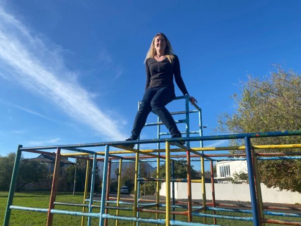 In a park, Lilley sits at the very top of a multicoloured jungle gym consisting of metal poles in a geometric structure, as if looking down from a throne. She is wearing a black long sleeved top with a v-neckline and dark blue jeans. She is also wearing a long chain around her neck with a symbol of a three-headed dragon on the disc hanging off it. Some of her long blonde hair is over her left shoulder and the rest flows down her back.