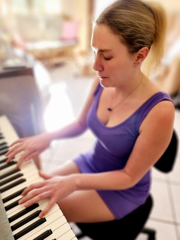 Lilley sits playing at a musical keyboard, deep in concentration. She is wearing a short purple dress and her blonde hair is held up in a clip at the back. There is a black obsidian arrow hanging from a silver chain around her neck. In the background there is a blurred bokeh of Lilley's apartment.