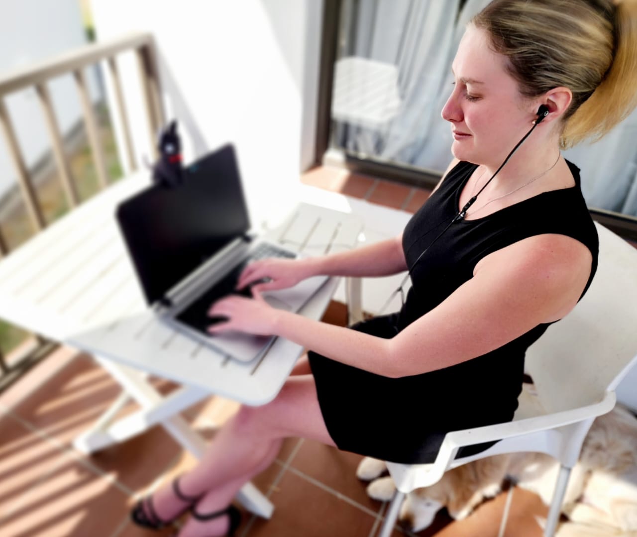 Lilley sits at a table, typing on a laptop with an earphone in one ear. She is wearing a tailored business-like black dress and black high heals, her long blonde hair is worn up in a hair clip, and she is wearing light makeup. There is a black cat plushy sitting on top of her laptop screen, and Lilley's guide dog Teska's face is just poking out from underneath her chair.